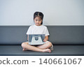 Young girl sitting cross-legged on a sofa using a tablet device with a neutral background, indoor setting, casual attire, focused expression, healthy lifestyle 140661067