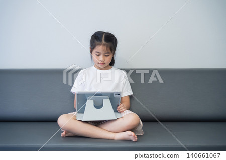 Young girl sitting cross-legged on a sofa using a tablet device with a neutral background, indoor setting, casual attire, focused expression, healthy lifestyle 140661067