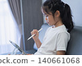 Young girl sitting on a sofa using a digital tablet, holding a white stylus, focused on screen, natural lighting, casual clothing, home interior, modern lifestyle 140661068
