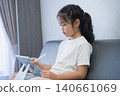 Young Girl Using Tablet Device Sitting on Sofa In Living Room Playing Educational Game or Watching Video With Pen in Hand Indoors 140661069