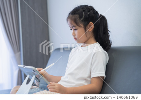 Young Girl Using Tablet Device Sitting on Sofa In Living Room Playing Educational Game or Watching Video With Pen in Hand Indoors Young Girl Using Tablet Device Sitting on Sofa In Living Room Playing Educational Game or Watching Video With Pen in Hand Indoors 140661069