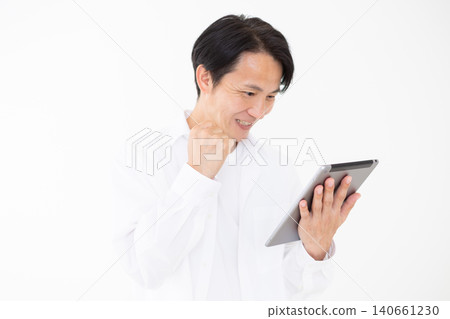 Portrait of a middle-aged man in a white shirt making a triumphant gesture while looking at a tablet, against a white background. Portrait of a middle-aged man in a white shirt making a triumphant gesture while looking at a tablet, against a white background. 140661230