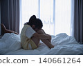 Young woman sitting alone on bed with legs drawn up, embracing her knees, in a dimly lit bedroom with large window and curtains, feeling sad or reflective 140661264