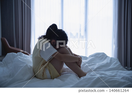 Young woman sitting alone on bed with legs drawn up, embracing her knees, in a dimly lit bedroom with large window and curtains, feeling sad or reflective Young woman sitting alone on bed with legs drawn up, embracing her knees, in a dimly lit bedroom with large window and curtains, feeling sad or reflective 140661264