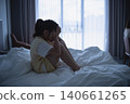 Young girl sitting on bed in dark room with her hands on her head, experiencing stress or sadness, near large window with closed curtains, gloomy atmosphere 140661265