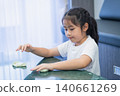 Young girl in casual clothing concentrating while playing with slime on a table in a modern room with curtains and a blue chair in the background 140661269