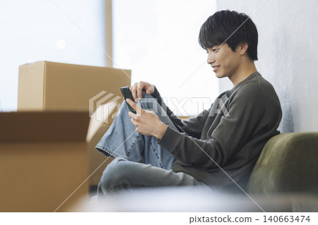 一名年輕男子被紙箱包圍,正在操作智慧型手機。 (畫面移動) 一名年輕男子被紙箱包圍,正在操作智慧型手機。 (畫面移動) 140663474