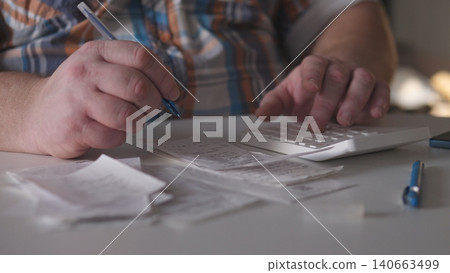 A person is sitting at a table, writing notes with a pen while using a calculator. Receipts are laid out on the table showing the busy work environment in an office setting. A person is sitting at a table, writing notes with a pen while using a calculator. Receipts are laid out on the table showing the busy work environment in an office setting. 140663499