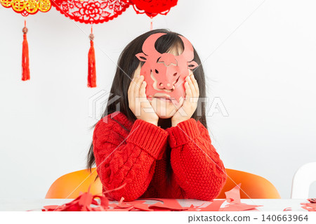 A little Asian girl wearing a bright red sweater is intently cutting red paper, preparing to welcome the Lunar New Year. A little Asian girl wearing a bright red sweater is intently cutting red paper, preparing to welcome the Lunar New Year. 140663964