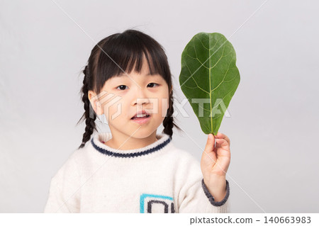 Little Asian girl covering half of her face with a large green leaf, concept of nature, environment and eco-friendly lifestyle. Little Asian girl covering half of her face with a large green leaf, concept of nature, environment and eco-friendly lifestyle. 140663983