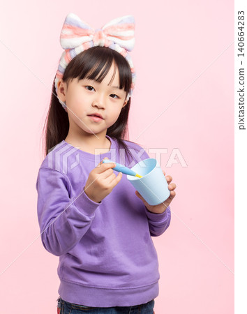 A studio portrait of an elementary-aged Asian girl with long dark hair, bangs, and a striped headband, wearing a purple sweatshirt, looking at the camera while holding a blue cup, posing on a pink A studio portrait of an elementary-aged Asian girl with long dark hair, bangs, and a striped headband, wearing a purple sweatshirt, looking at the camera while holding a blue cup, posing on a pink 140664283