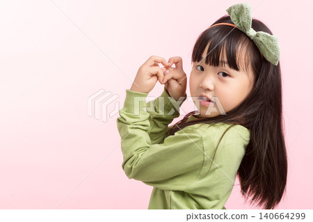 A side view portrait of a young Asian girl with long dark hair and a green bow headband, wearing a green sweatshirt, making a heart shape gesture with her hands near her face against a pink studio 140664299