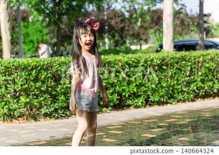 A joyful young girl runs playfully on a wet paved park path in bright sunlight. A joyful young girl runs playfully on a wet paved park path in bright sunlight. 140664324