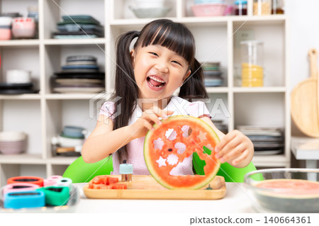 Laughing young Asian girl proudly displaying large watermelon slice with star cutouts at kitchen table Laughing young Asian girl proudly displaying large watermelon slice with star cutouts at kitchen table 140664361