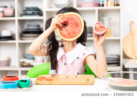 Playful young Asian girl covering one eye with watermelon slice while holding heart shaped toy in other hand Playful young Asian girl covering one eye with watermelon slice while holding heart shaped toy in other hand 140664363