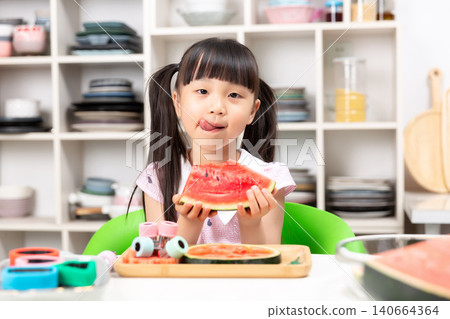 Cheeky young Asian girl while holding watermelon slice at kitchen table with bright smile Cheeky young Asian girl while holding watermelon slice at kitchen table with bright smile 140664364