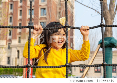 Joyful little girl smiles excitedly while playing inside a rope climbing net at an outdoor playground. Joyful little girl smiles excitedly while playing inside a rope climbing net at an outdoor playground. 140665219