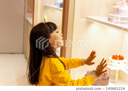Curious little girl looks through a glass display case at beautiful multi-layered celebration cakes. Curious little girl looks through a glass display case at beautiful multi-layered celebration cakes. 140665224