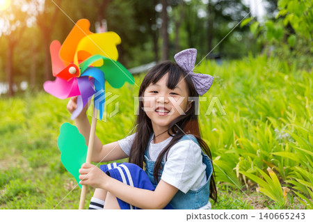Happy Young East Asian Girl with Purple Bow Sitting in Sunny Green Meadow Holding Colorful Rainbow Pinwheel with Joyful Smile and Bright Summer Vibes. Happy Young East Asian Girl with Purple Bow Sitting in Sunny Green Meadow Holding Colorful Rainbow Pinwheel with Joyful Smile and Bright Summer Vibes. 140665243