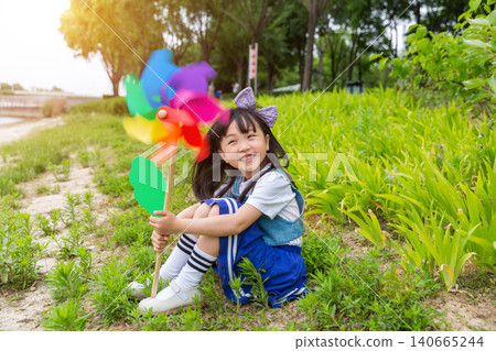 Happy Young East Asian Girl with Purple Bow Sitting in Sunny Green Meadow Holding Colorful Rainbow Pinwheel with Joyful Smile and Bright Summer Vibes. 140665244