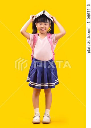 A cute Asian female student stands against a yellow background with a book on her head, happily and cheerfully welcoming the new semester. A cute Asian female student stands against a yellow background with a book on her head, happily and cheerfully welcoming the new semester. 140665246