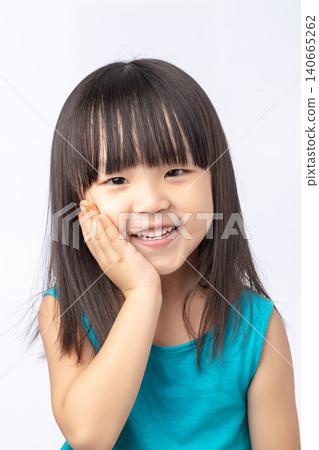 Close-up portrait of a cheerful little Asian girl in a blue dress, smiling with hand on cheek. Close-up portrait of a cheerful little Asian girl in a blue dress, smiling with hand on cheek. 140665262