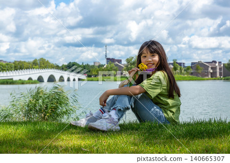 A happy young girl sitting on the green grass by a lake while holding yellow wild flowers on a cloudy day. A happy young girl sitting on the green grass by a lake while holding yellow wild flowers on a cloudy day. 140665307