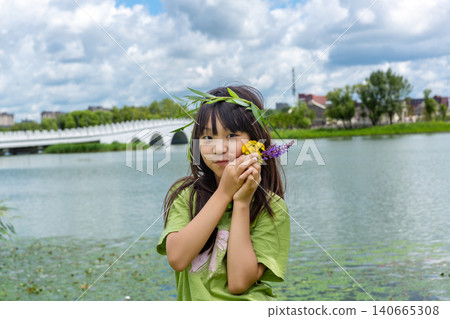A young girl wearing a handmade willow garland and holding wildflowers smiles near a scenic riverbank. A young girl wearing a handmade willow garland and holding wildflowers smiles near a scenic riverbank. 140665308