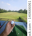 A first-person perspective shot shows a person holding a white golf ball over a green practice mat, with a vast driving range field and distant trees visible under a clear sky. 140666061