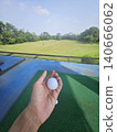 A first-person perspective shot shows a person holding a white golf ball over a green practice mat, with a vast driving range field and distant trees visible under a clear sky. 140666062