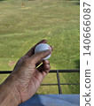 A first-person perspective shot shows a person holding a white golf ball over a green practice mat, with a vast driving range field and distant trees visible under a clear sky. 140666087