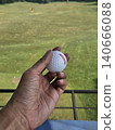 A first-person perspective shot shows a person holding a white golf ball over a green practice mat, with a vast driving range field and distant trees visible under a clear sky. 140666088