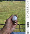 A first-person perspective shot shows a person holding a white golf ball over a green practice mat, with a vast driving range field and distant trees visible under a clear sky. 140666094