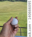 A first-person perspective shot shows a person holding a white golf ball over a green practice mat, with a vast driving range field and distant trees visible under a clear sky. 140666095