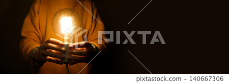 Light bulb glowing in hands with copy space on dark background. Horizontal panoramic banner of person holding bright incandescent lamp, creativity, inspiration and energy crisis concept 140667306