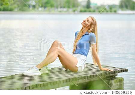 Woman sitting on pier by lake enjoying sunny day with water and trees in background Woman sitting on pier by lake enjoying sunny day with water and trees in background 140669139