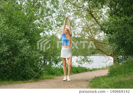 Young woman in sporty outfit stretches by path near water during daytime in green park Young woman in sporty outfit stretches by path near water during daytime in green park 140669142