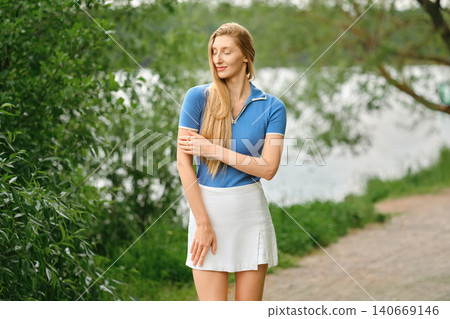 Woman walking by water in blue top and white skirt on sunny day near trees and path Woman walking by water in blue top and white skirt on sunny day near trees and path 140669146