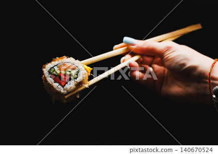 Sushi roll held with chopsticks above a dark background showing the colorful ingredients inside the roll at a dining table 140670524
