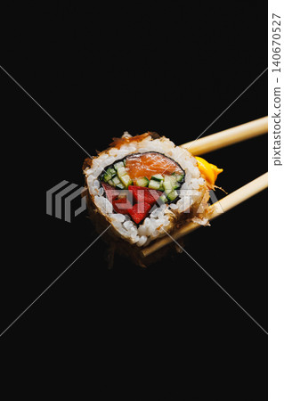Sushi roll with fresh ingredients held by chopsticks on a black background in a kitchen setting Sushi roll with fresh ingredients held by chopsticks on a black background in a kitchen setting 140670527