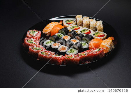 A variety of sushi rolls and sashimi served on a black plate in a restaurant setting during dinner time A variety of sushi rolls and sashimi served on a black plate in a restaurant setting during dinner time 140670536