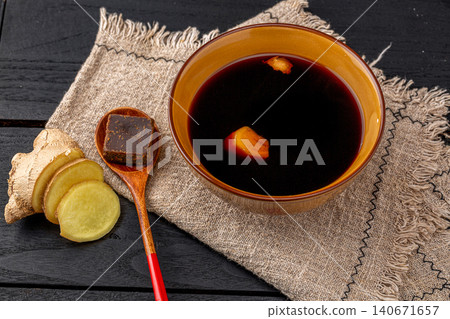 a bowl of hot ginger tea with brown sugar cubes and fresh ginger slices on a rustic burlap mat, asian herbal health drink. a bowl of hot ginger tea with brown sugar cubes and fresh ginger slices on a rustic burlap mat, asian herbal health drink. 140671657