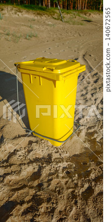 Yellow waste bin positioned on sandy beach with natural vegetation and trees visible in the background under bright sunlight Yellow waste bin positioned on sandy beach with natural vegetation and trees visible in the background under bright sunlight 140672565