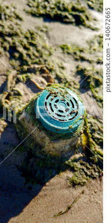 Blue circular drainage cover partially submerged in sand and seaweed on a beach, with sunlight illuminating the surrounding area and textures of the sand Blue circular drainage cover partially submerged in sand and seaweed on a beach, with sunlight illuminating the surrounding area and textures of the sand 140672665