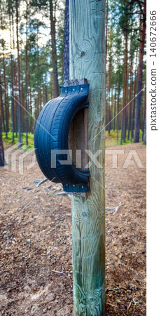 Black rubber tire mounted on a wooden post in a forest setting, surrounded by tall trees and a natural ground cover of leaves and pine needles Black rubber tire mounted on a wooden post in a forest setting, surrounded by tall trees and a natural ground cover of leaves and pine needles 140672666