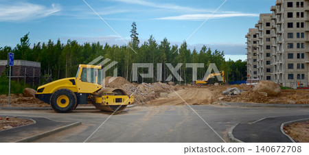 Yellow road roller and excavator on construction site with unfinished building and forest in the background under a clear blue sky 140672708