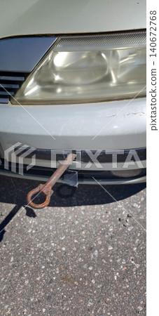 Rusty tow hook protruding from the front bumper of a silver sedan parked on a paved surface under bright sunlight 140672768