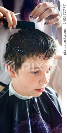Young boy receiving a haircut at a salon, stylist using a comb to style hair while wearing a black cape, with soft lighting illuminating the scene Young boy receiving a haircut at a salon, stylist using a comb to style hair while wearing a black cape, with soft lighting illuminating the scene 140672777