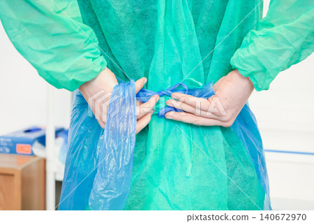 Medical worker ties a blue plastic disposable apron over a green protective gown in a clinical setting to ensure hygiene compliance and safety protocols during a daily hospital shift Medical worker ties a blue plastic disposable apron over a green protective gown in a clinical setting to ensure hygiene compliance and safety protocols during a daily hospital shift 140672970