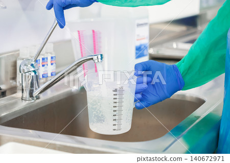 A laboratory worker in protective gear fills a plastic measuring cup with water at a stainless steel sink for precise liquid preparation tasks within a clean clinical facility A laboratory worker in protective gear fills a plastic measuring cup with water at a stainless steel sink for precise liquid preparation tasks within a clean clinical facility 140672971